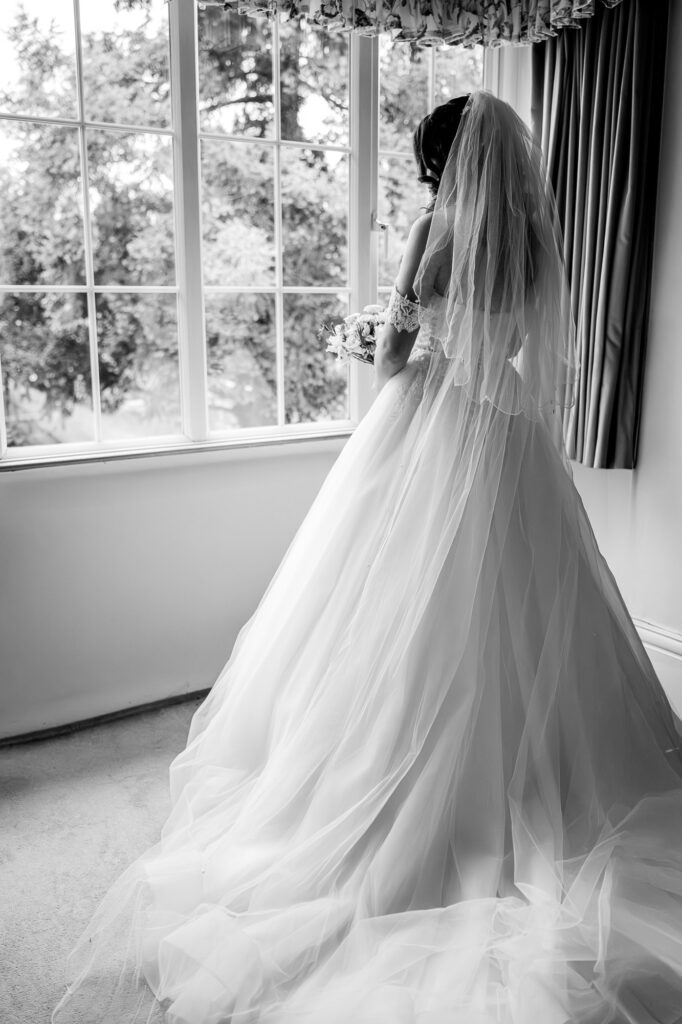 A bride in a flowing wedding gown and veil stands by a window, looking outside and holding a bouquet, with soft natural light streaming in. Black and white photograph.