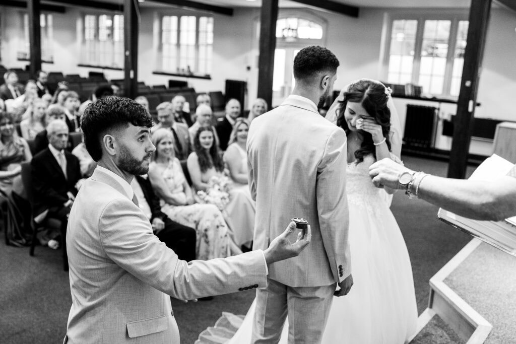 A man kneels and presents wedding rings during a ceremony, while a bride wipes her eyes and a groom stands beside her. Guests watch the emotional moment in a church setting.