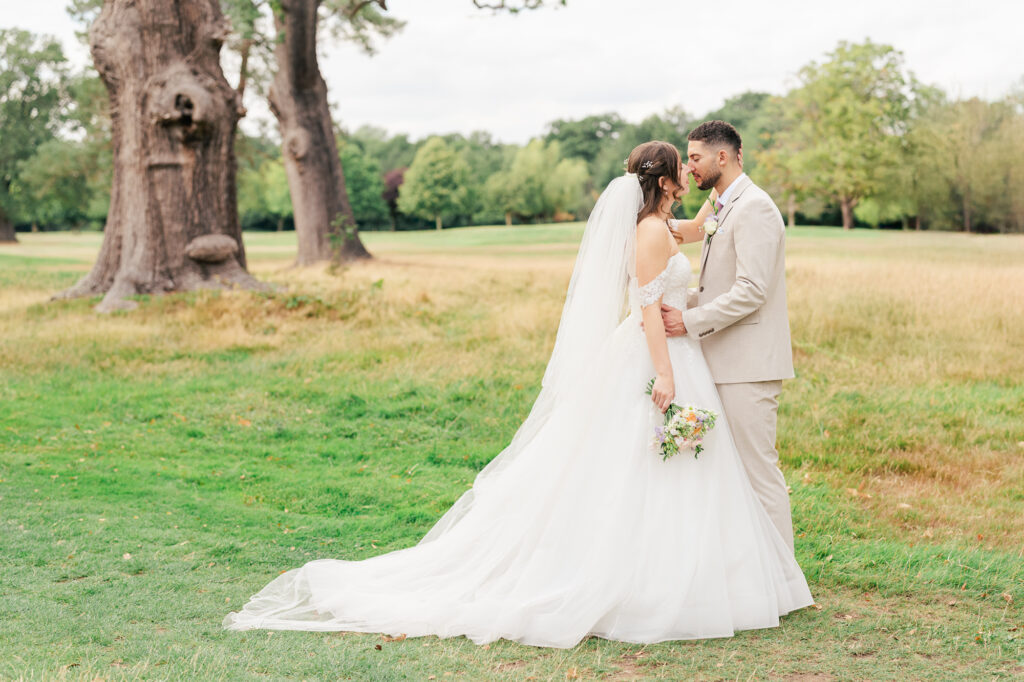 A bride and groom stand facing each other outdoors on grass, embracing and smiling. The bride wears a white dress and veil, holding a bouquet. The groom wears a light tan suit. Trees and a field are in the background.