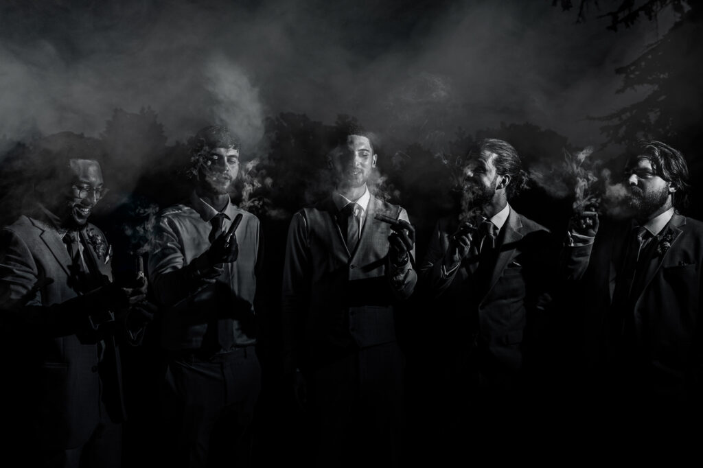 Five men in suits stand together at night, each holding and smoking a cigar. Smoke partially obscures their faces and drifts through the air, creating a dramatic and moody atmosphere. Trees are faintly visible in the background.
