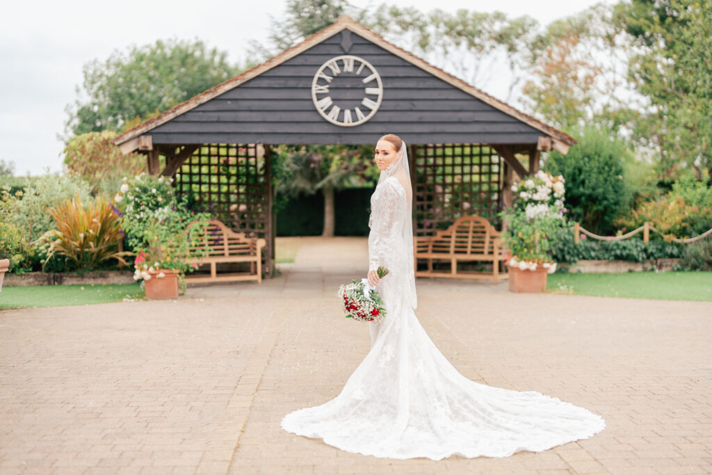 A bride in a long, white lace gown and veil stands holding a bouquet in front of a wooden gazebo with a large clock, surrounded by greenery and benches.