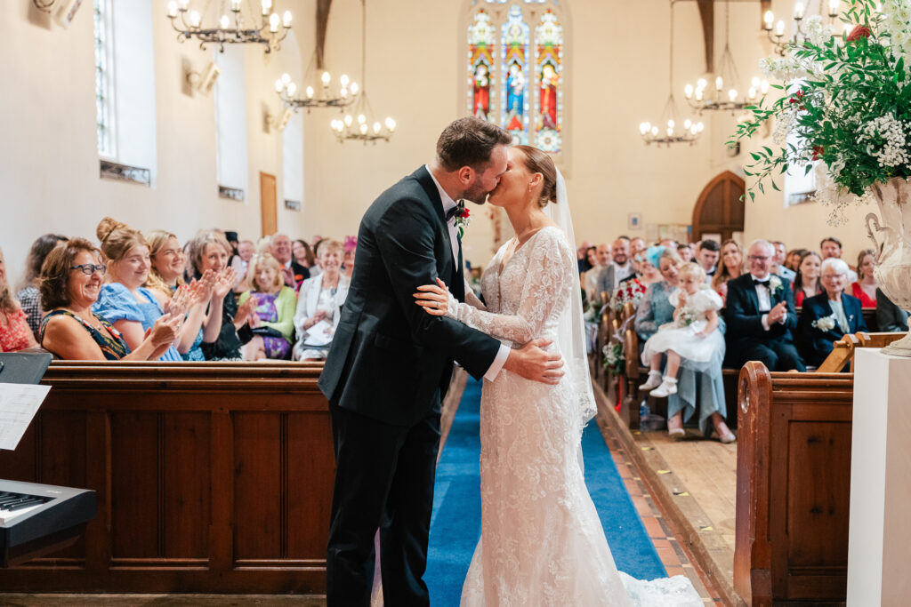 A bride and groom kiss at the altar of a church filled with seated guests, who smile and clap. The bride wears a lace gown and veil, and the groom wears a dark suit. Stained glass windows are visible in the background.