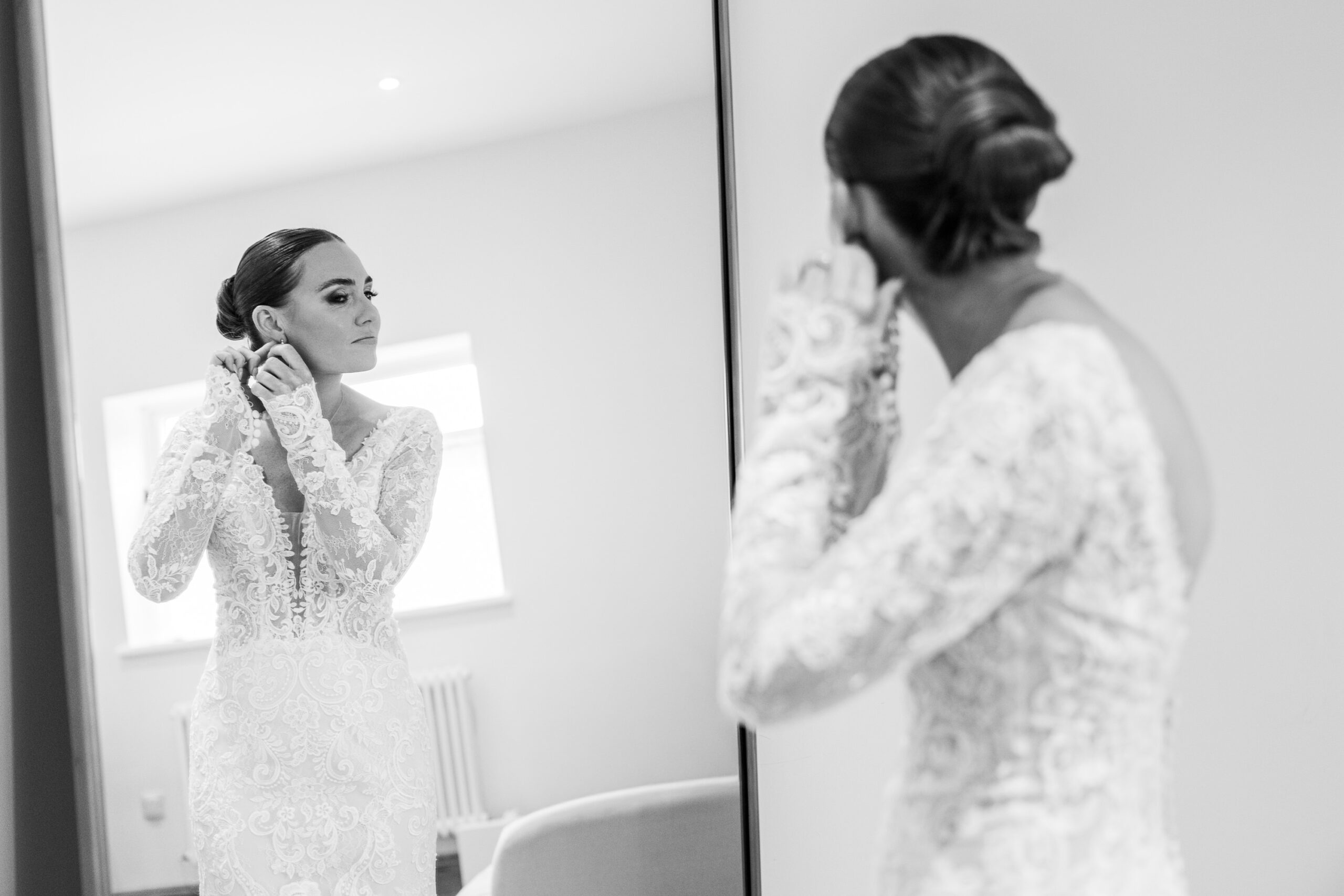 3934336342 A woman in a lace wedding dress stands in front of a mirror, putting on an earring. Her hair is styled in a low bun and she appears focused, preparing for her wedding day. The image is in black and white.