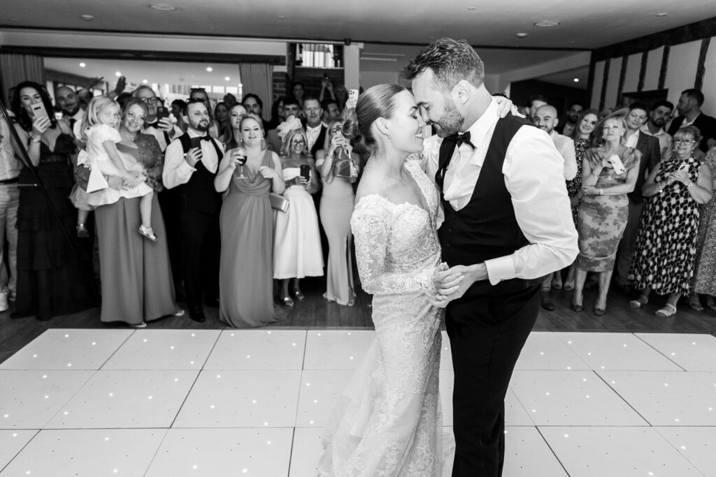 A bride and groom share their first dance on a lit dance floor, surrounded by smiling guests who watch and take photos. The couple gazes at each other, and the venue is bright and festive.
