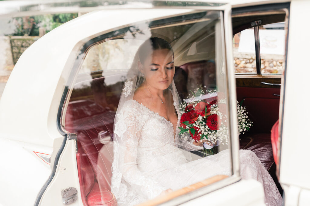 A bride in a lace wedding dress and veil sits in the backseat of a vintage car, holding a bouquet of red and white flowers, looking down with a serene expression.