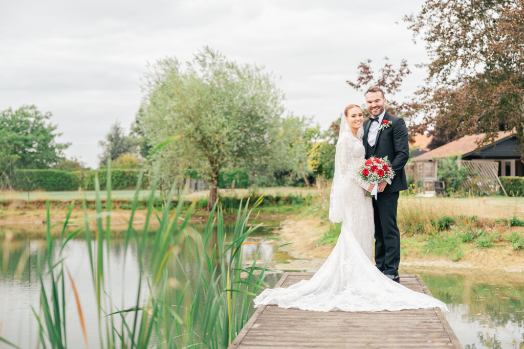 A bride and groom stand together on a wooden dock by a pond, smiling at the camera. The bride wears a white lace gown and holds a red bouquet; the groom is in a black tuxedo. Greenery and trees surround them.