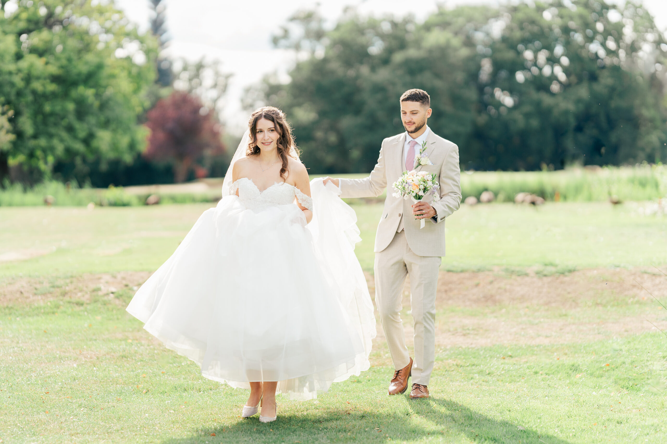 KF020825C-467 A bride in a white gown walks on grass, smiling, while a groom in a tan suit follows behind her holding a bouquet. They are outdoors in a sunny, green park setting.