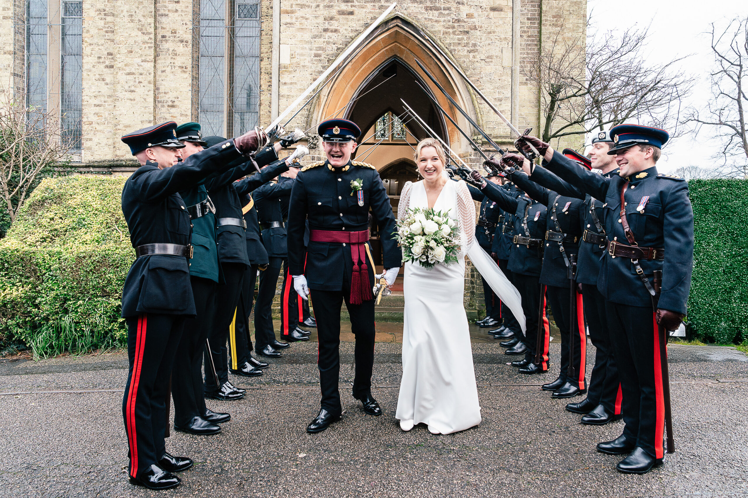 3673688123 A bride and groom walk under an archway of raised swords held by uniformed soldiers outside a stone church, smiling and holding hands. The bride carries a bouquet of white flowers.