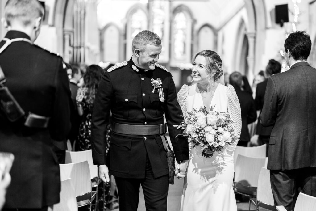A newly married couple walks down the aisle in a church, smiling at each other and holding hands. The groom wears a military uniform and the bride holds a bouquet, with guests seated on either side. The scene is in black and white.