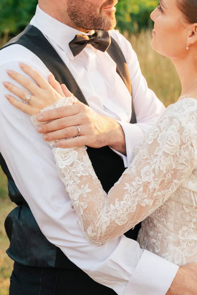 A bride and groom embrace outdoors. The bride wears a lace wedding dress and has her hand on the groom's chest, showing her wedding ring. The groom wears a black vest and bow tie. Their faces are close, sharing an intimate moment.
