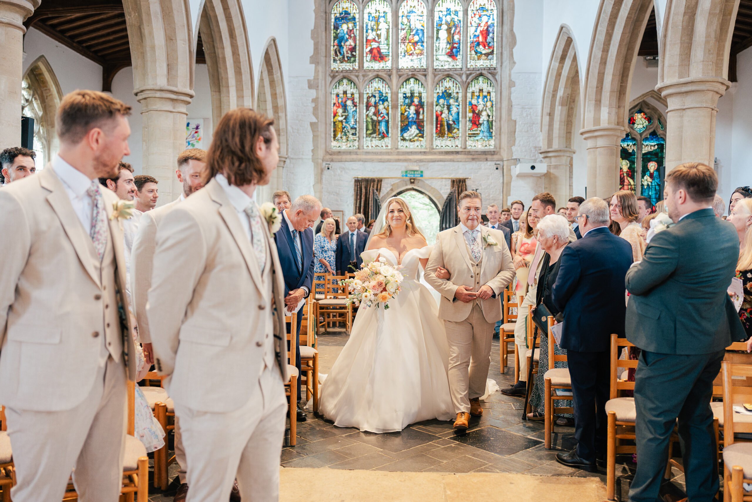 A bride in a white gown walks down the aisle with two men in beige suits, surrounded by guests seated in a church with stained glass windows and high arches.