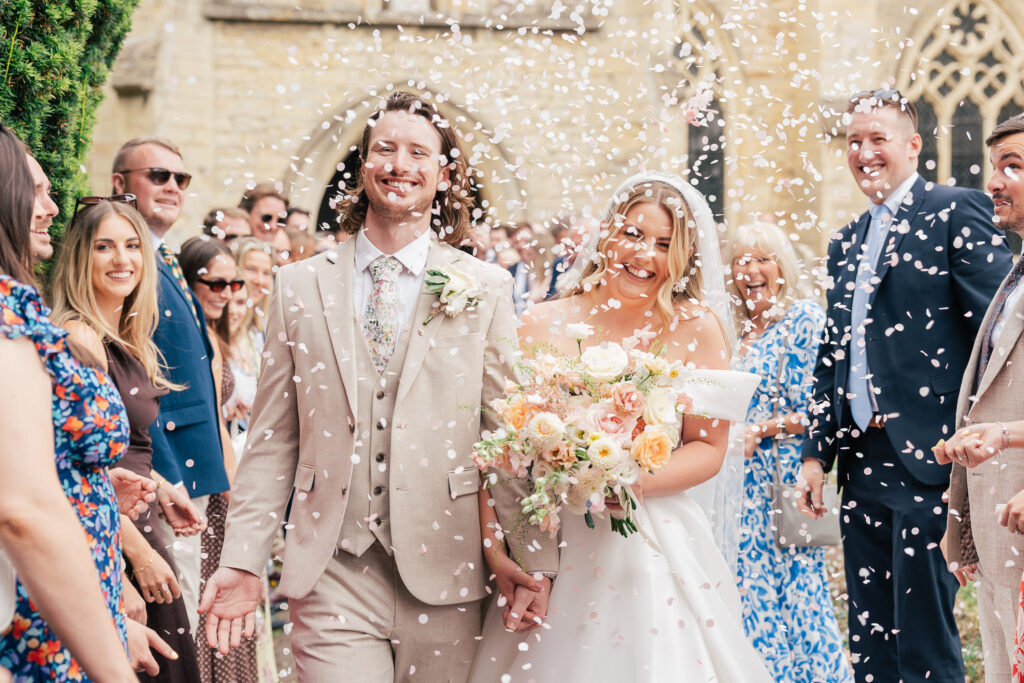 A newly married couple smiling and walking outside a church as guests throw confetti. The groom wears a beige suit and the bride holds a bouquet of flowers, surrounded by cheering friends and family.