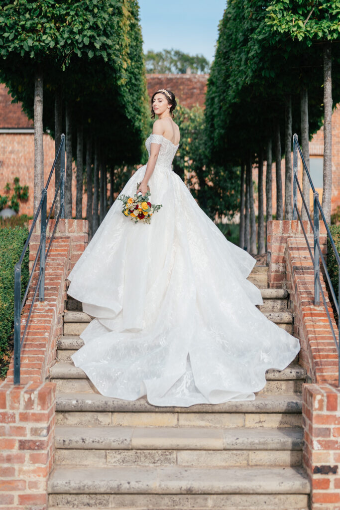 A bride in an elegant white gown stands on stone steps between manicured trees, holding a bouquet of yellow and orange flowers, looking over her shoulder towards the camera.