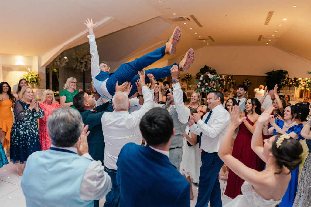 A group of people at a wedding reception lift a man in a blue suit into the air while smiling and cheering. Guests in colorful attire surround them, clapping and celebrating in a decorated indoor venue.