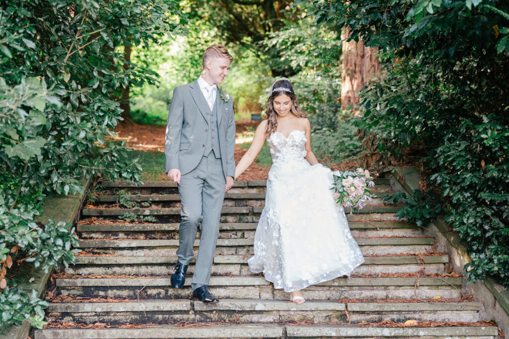 A bride in a white floral gown and a groom in a light grey suit walk hand-in-hand down outdoor stone steps surrounded by lush greenery. The bride holds a bouquet and both are smiling happily.