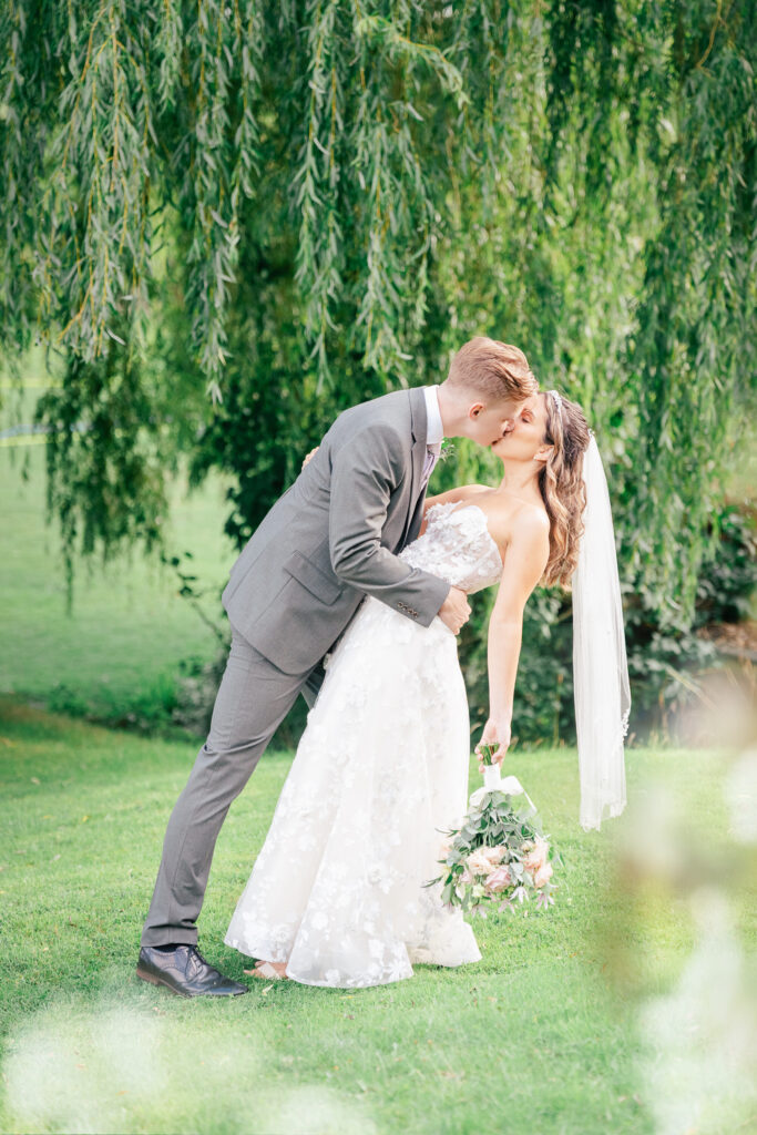 A groom in a gray suit dips and kisses a bride in a strapless white gown and veil, holding a bouquet, on lush green grass under leafy trees in a sunlit outdoor setting.