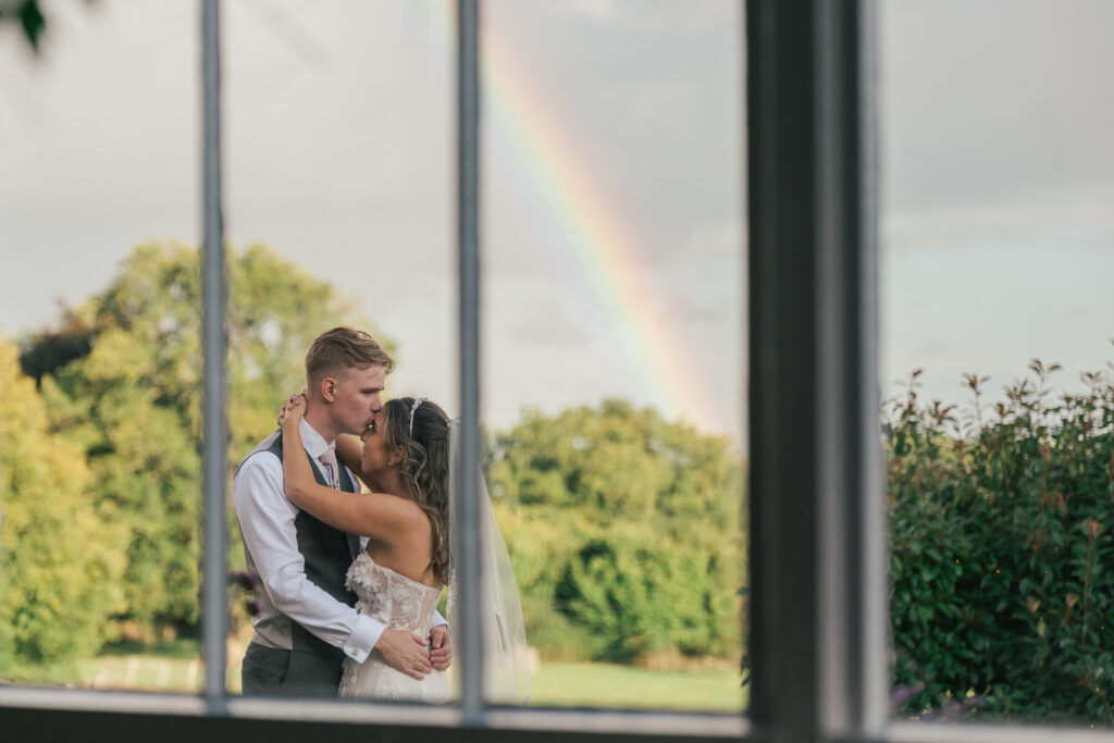 A bride and groom share a kiss outdoors, framed by a window, with a vibrant rainbow and lush green trees in the background.