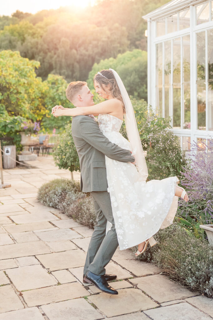 A groom in a grey suit lifts his smiling bride in a white floral dress and veil outside, surrounded by greenery and bathed in warm sunlight near a glass greenhouse.