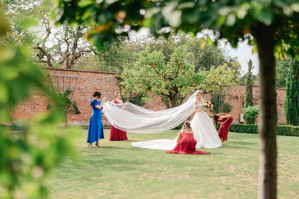 A bride in a garden stands holding a bouquet while three bridesmaids in red dresses and a woman in blue adjust her long veil, surrounded by trees, grass, and a brick wall.