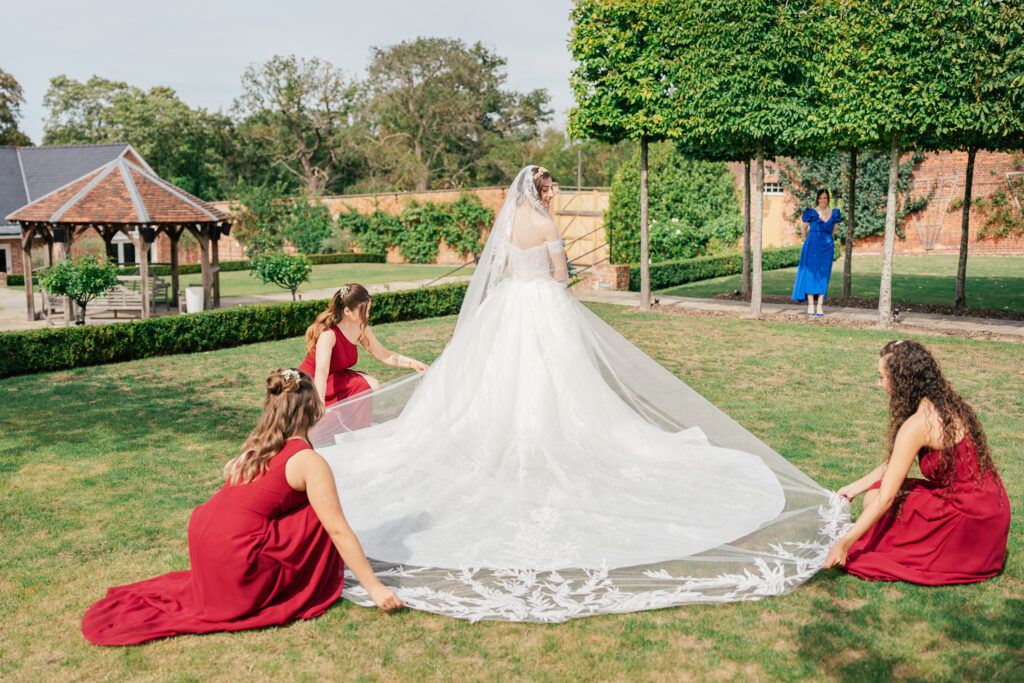 A bride in a white gown stands on grass while three bridesmaids in red dresses arrange her long train. In the background, a woman in blue waves near trees and a brick wall. The setting appears to be an outdoor garden.