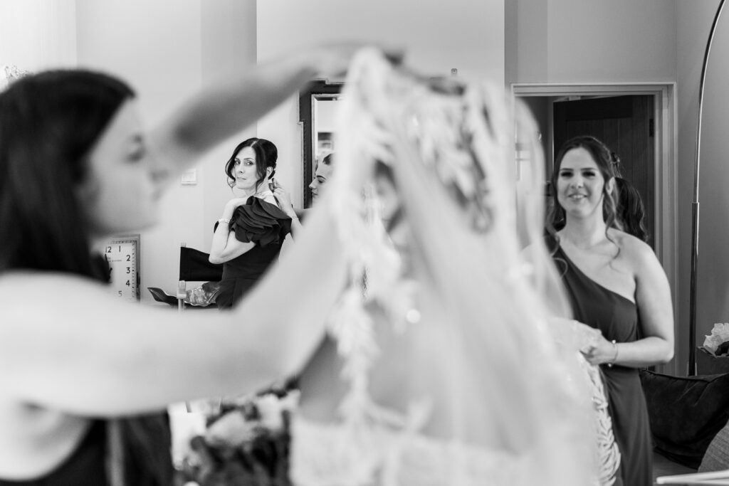 Black and white photo of women preparing for a wedding; one holds a dress in the foreground, while three others, wearing dresses, are seen in the background getting ready and smiling.