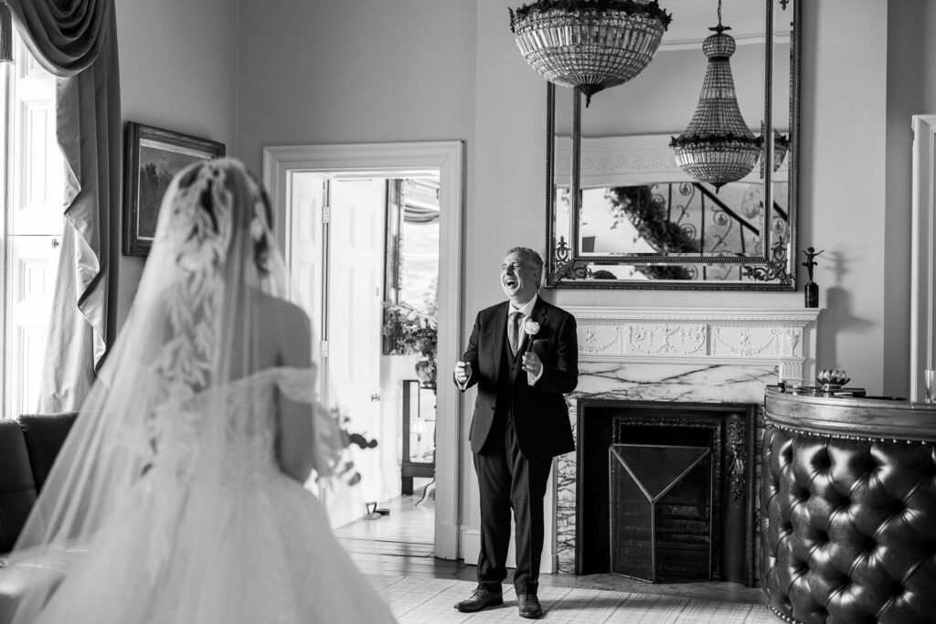 A bride in a wedding dress stands facing an older man in a suit, who smiles joyfully with open arms in an elegant room with chandeliers and a marble fireplace.
