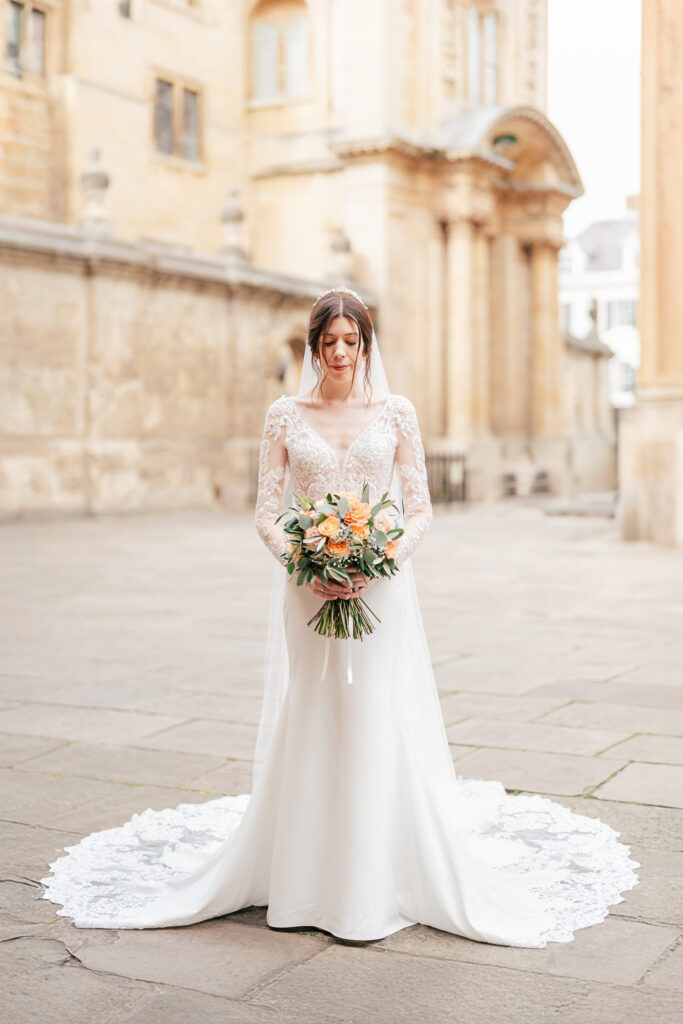 A bride in a long-sleeved lace wedding dress with a train stands outdoors on a stone pathway, holding a bouquet of yellow and white flowers, with historic stone buildings in the background.