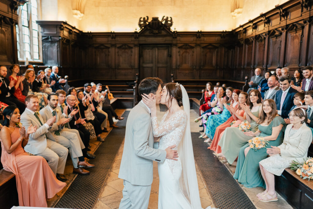 A bride and groom kiss at the altar in a wood-paneled room as guests seated on both sides smile, clap, and celebrate during a wedding ceremony.