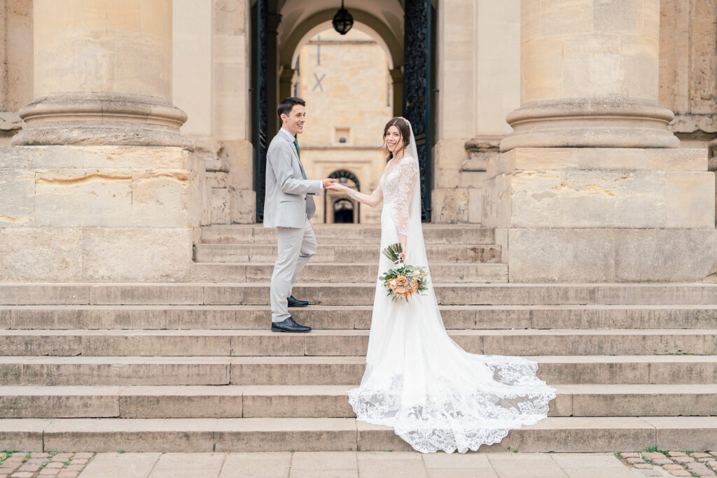 A bride in a long white dress holds a bouquet and the hand of a groom in a light grey suit as they stand on stone steps in front of a grand building with columns.