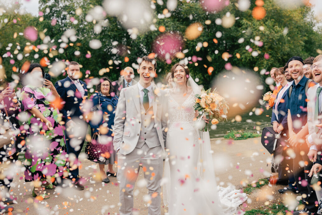 A newlywed couple smiles as they walk outdoors, surrounded by guests throwing colorful flower petals. The crowd is joyful, and the scene is filled with celebration and confetti in the air.