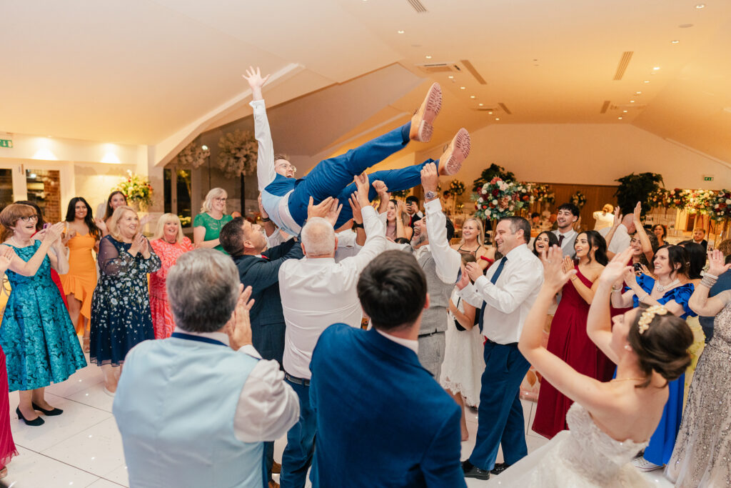 A joyous wedding party dances in a bright hall as a group lifts a man in a blue suit above their heads. Guests in colorful dresses and suits surround them, smiling and cheering with raised hands.