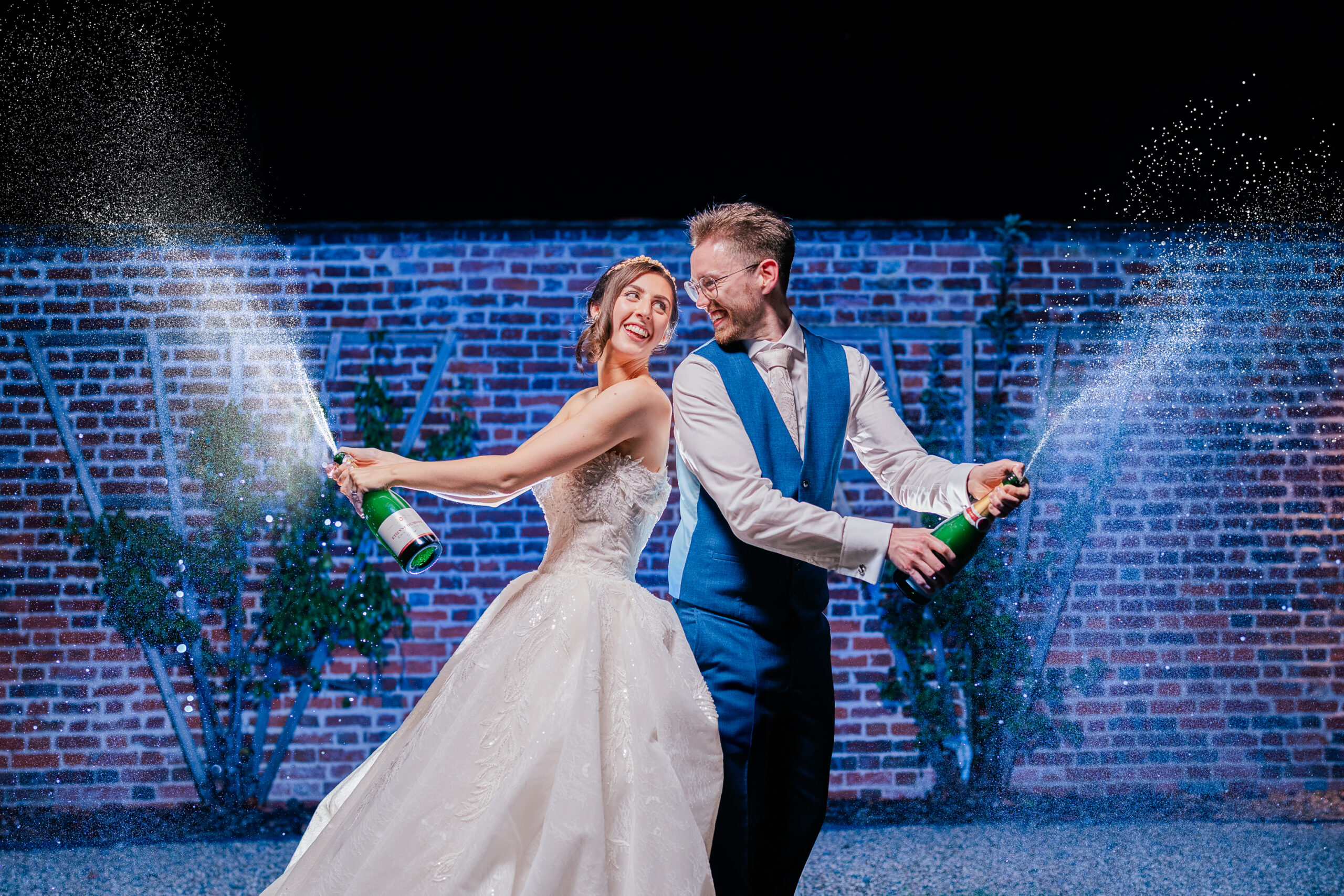 A bride in a white gown and a groom in a blue suit joyfully spray champagne from bottles, standing side by side at night in front of a brick wall decorated with greenery.