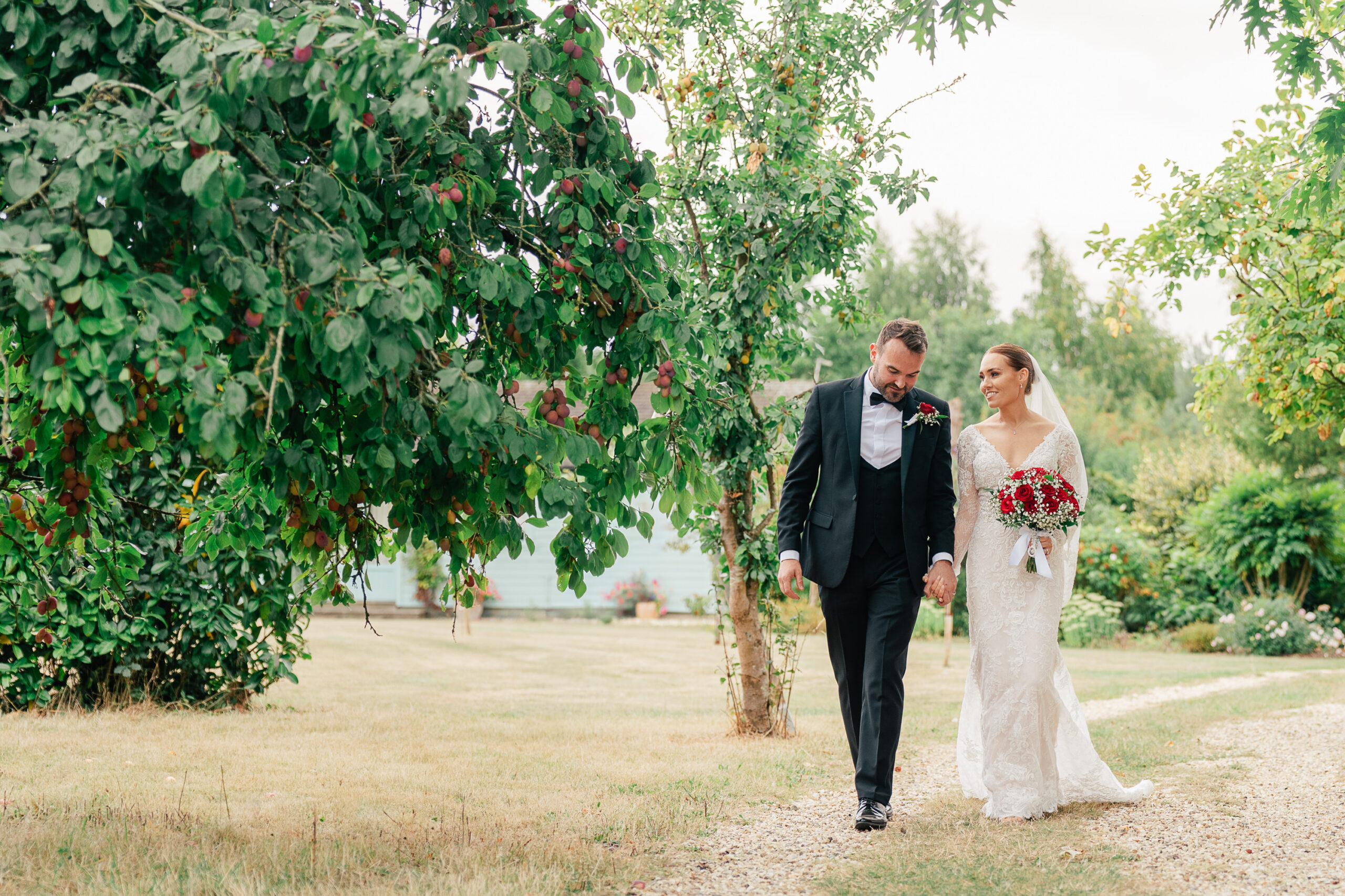A bride in a white gown and a groom in a black suit walk hand-in-hand down a garden path, surrounded by green trees and grass, with the bride holding a bouquet of red flowers.
