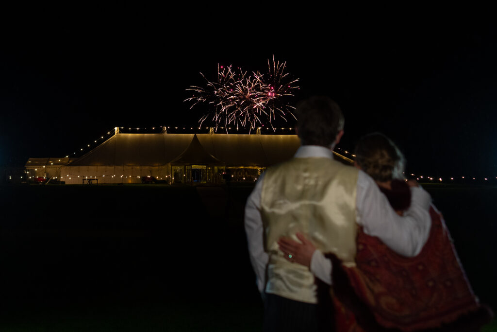 A couple stands arm in arm at night, watching colorful fireworks above a large, illuminated tent in the distance.