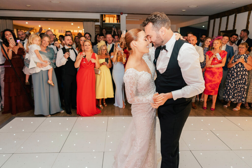 A bride and groom share their first dance at their wedding reception, smiling and holding each other closely while guests stand around them, watching and taking photos.