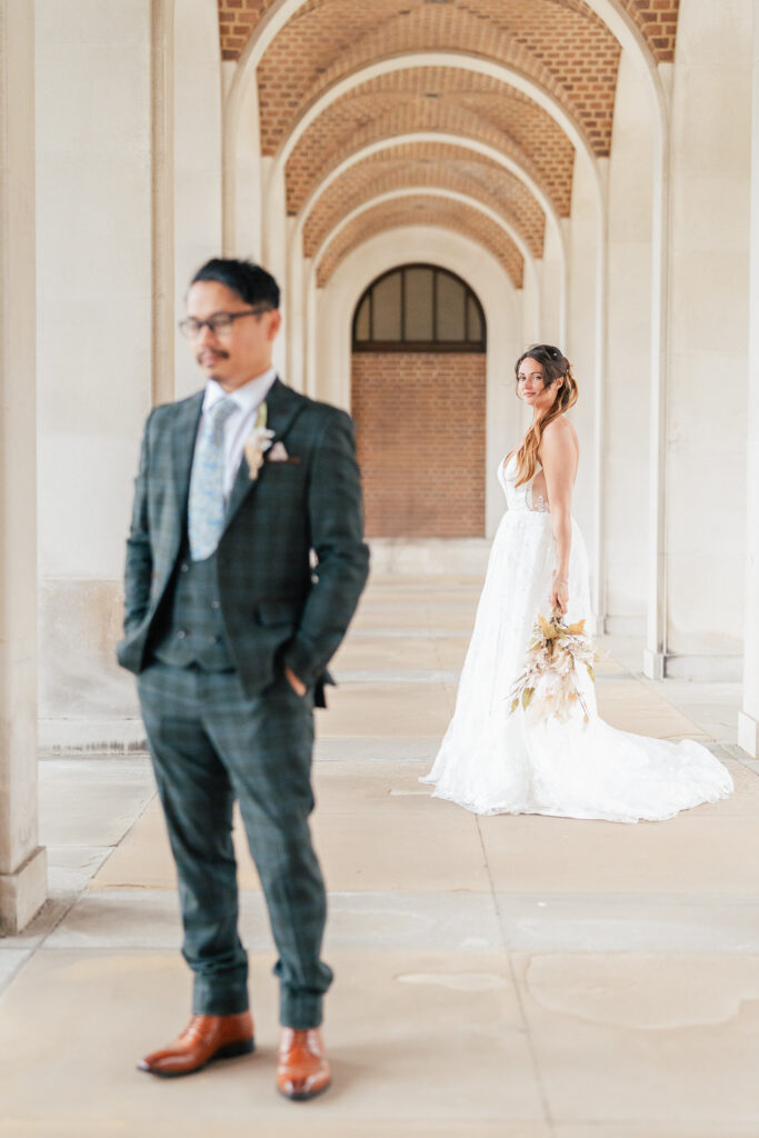 A bride in a white gown holding a bouquet stands in focus under an arched walkway, while a groom in a plaid suit stands out of focus in the foreground with hands in his pockets.