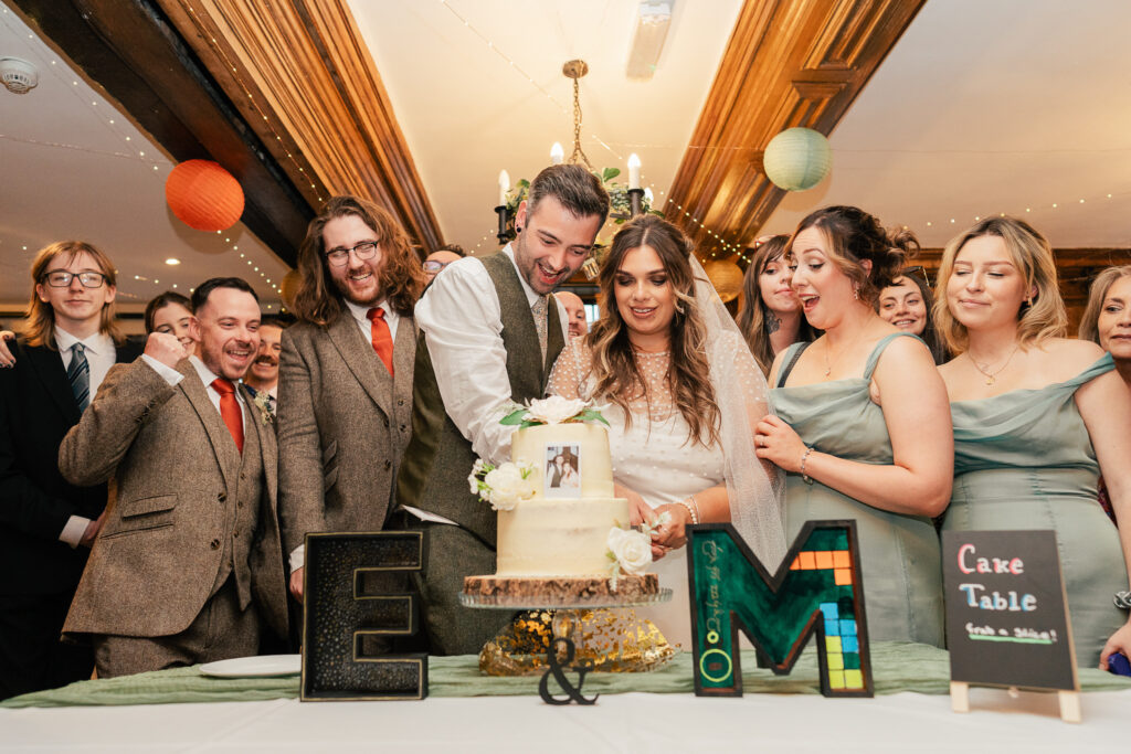 A bride and groom cut their wedding cake together, surrounded by smiling guests. The table has large decorative letters "E" and "M," flowers, and a small sign reading "Cake Table." The scene is festive and joyful.