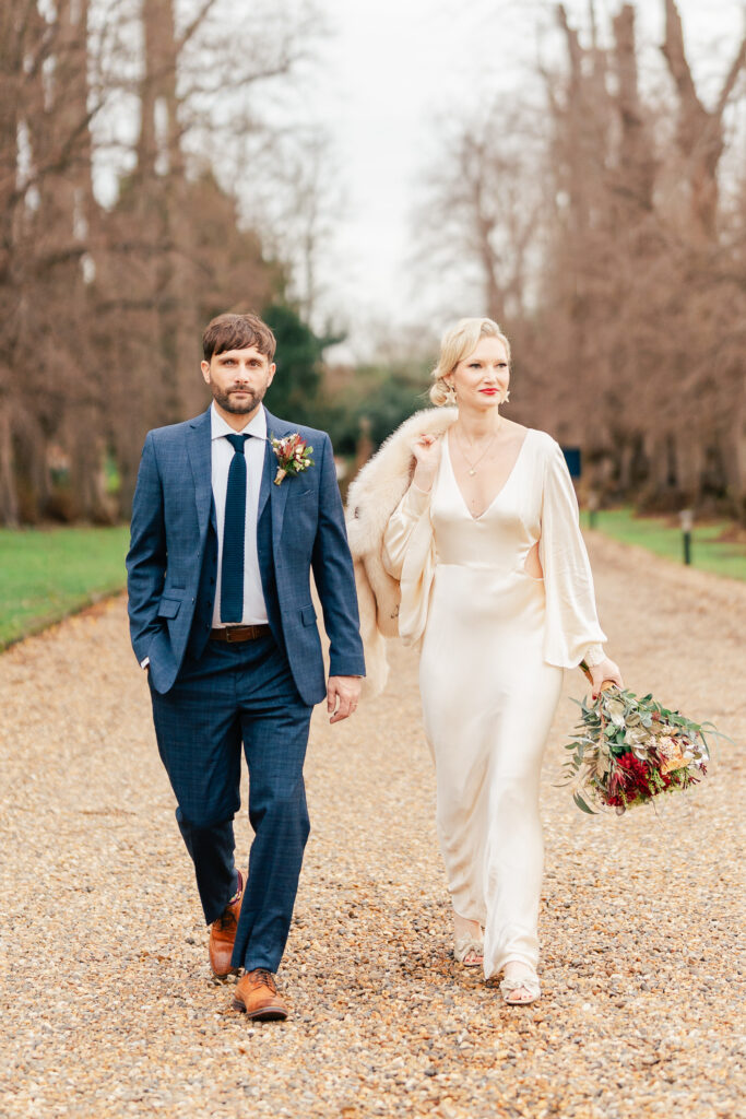 A man in a blue suit and a woman in a cream silk gown holding a bouquet walk side by side outdoors on a gravel path, surrounded by bare trees on a cloudy day.