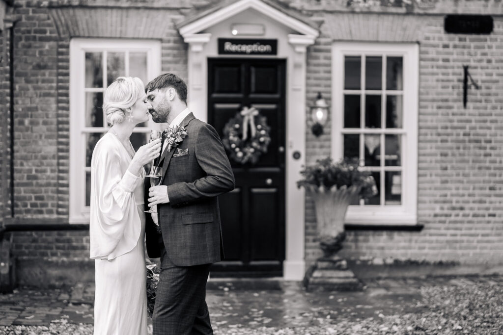 A bride and groom share an intimate moment outside a building with a "Reception" sign above the door. The bride holds flowers, and both are dressed formally. The scene is in black and white.