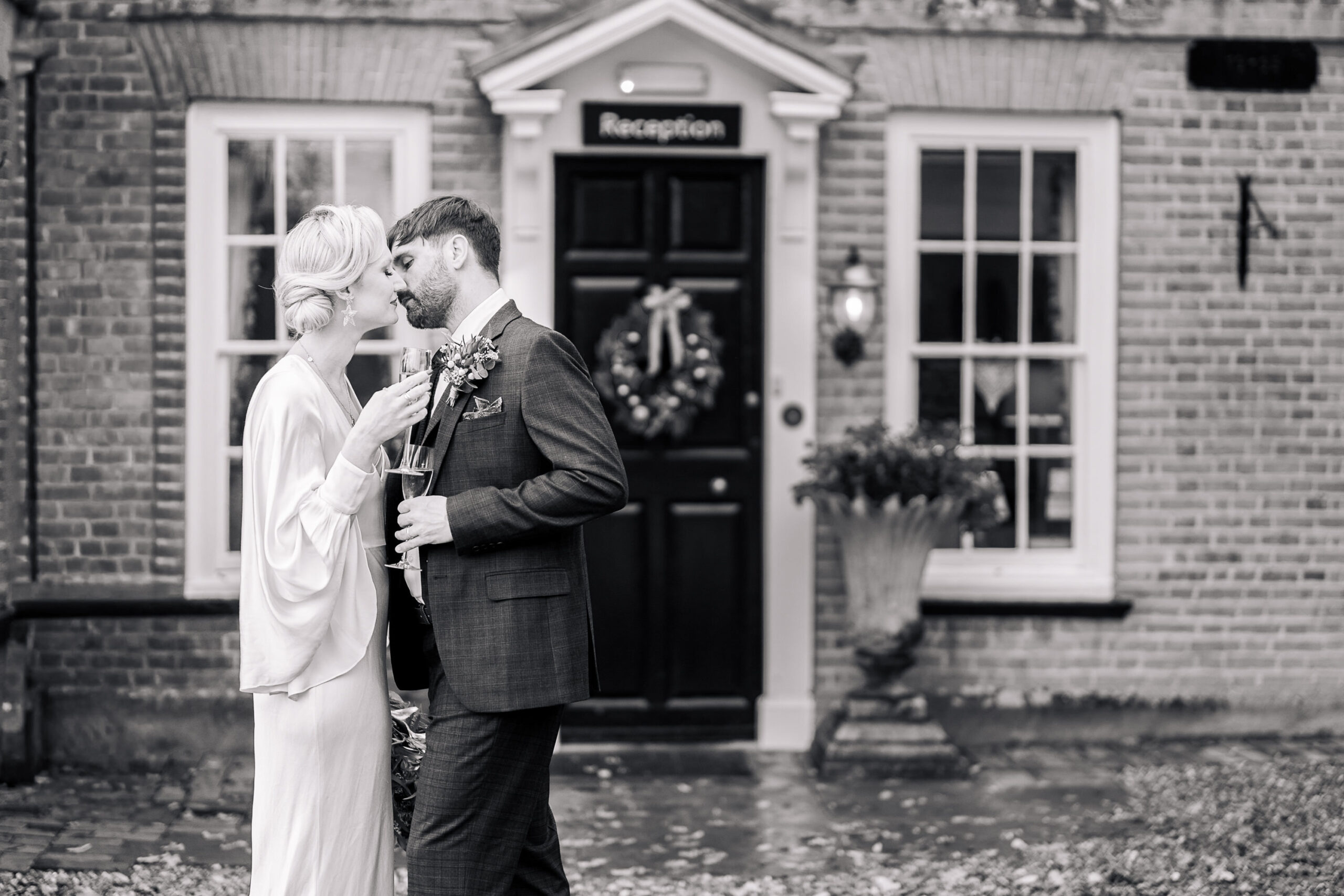 4095894961 A bride and groom share an intimate moment outside a building with a "Reception" sign above the door. The bride holds flowers, and both are dressed formally. The scene is in black and white.