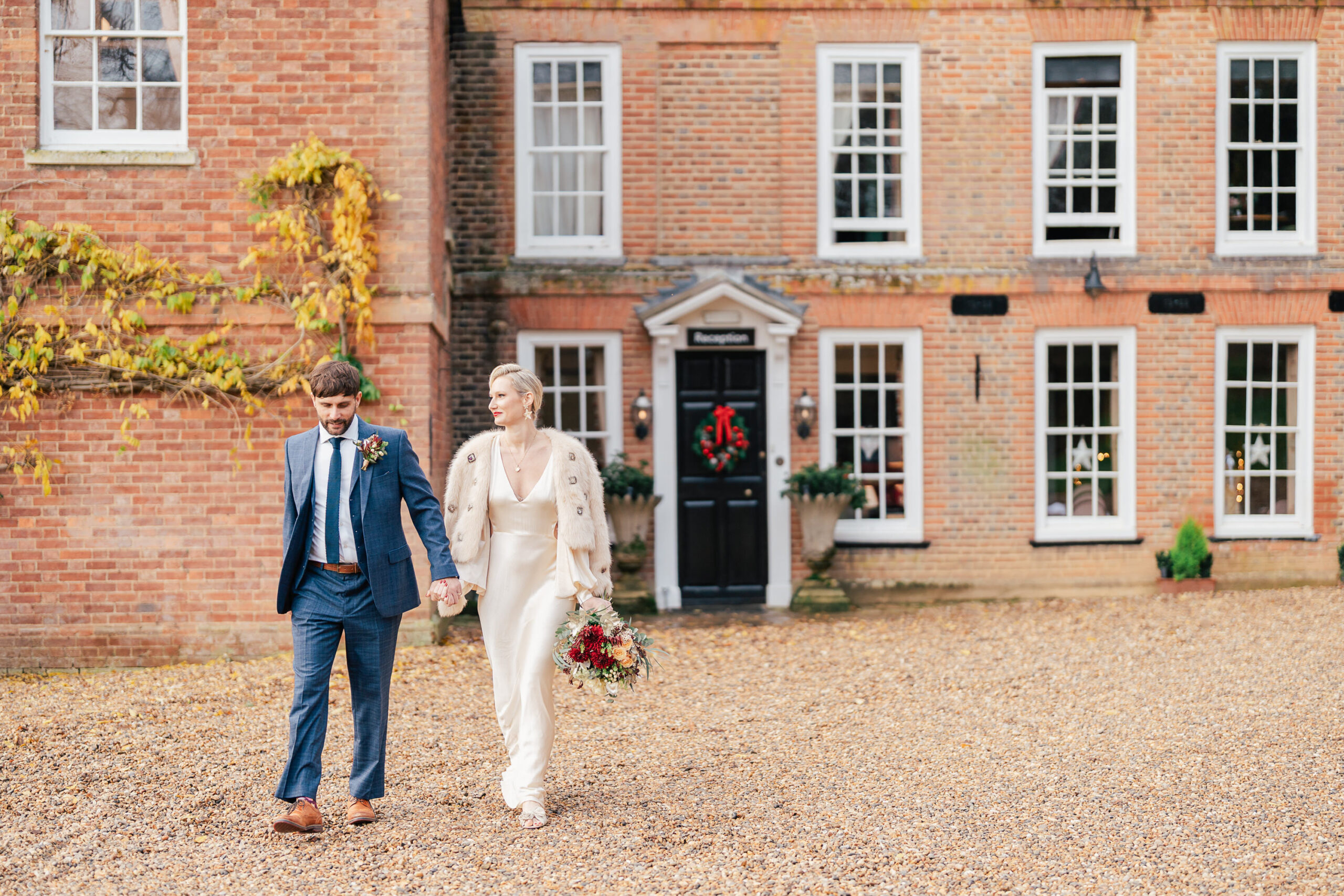 A bride in a white dress holding a bouquet walks hand in hand with a groom in a blue suit outside a brick building with white-trimmed windows and a black door decorated for the holidays.