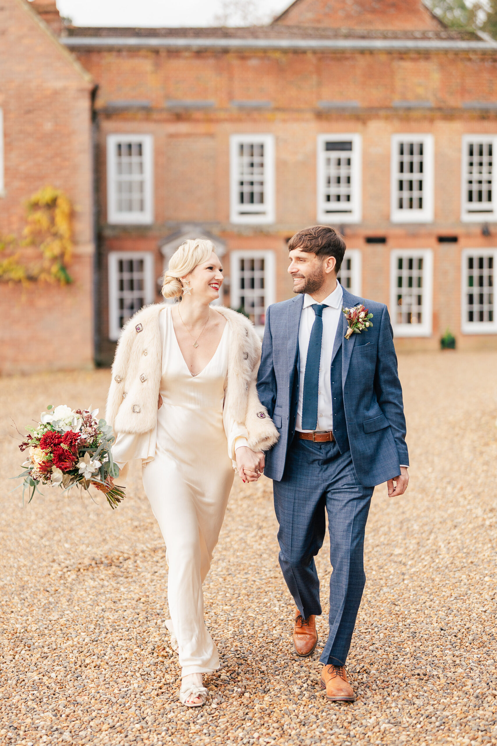 4095899132 A bride in a white dress and fur jacket holds a bouquet and walks hand-in-hand with a groom in a blue suit and tie. They smile at each other outside a stately brick building with tall windows.