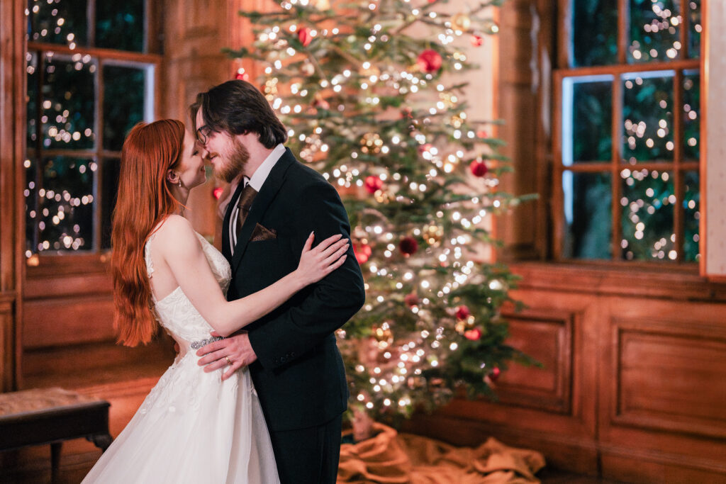 A bride and groom embrace and smile at each other in front of a decorated Christmas tree with twinkling lights, inside a warmly lit wooden-paneled room.