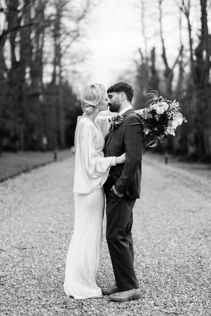 A black and white photo of a bride in a long gown and a groom in a suit standing close together on a gravel path, gazing into each other's eyes, with trees lining the background. The bride holds a bouquet.