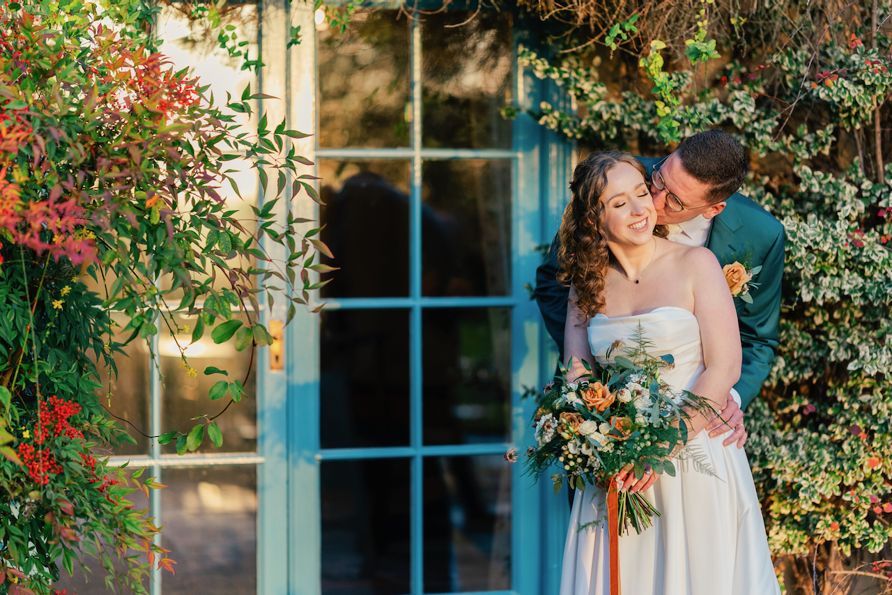 SouthFarm-5 (1) A groom in a green suit lovingly embraces and kisses his smiling bride, who holds a bouquet of flowers. They stand in front of a blue door framed by greenery and colorful flowers in warm sunlight.