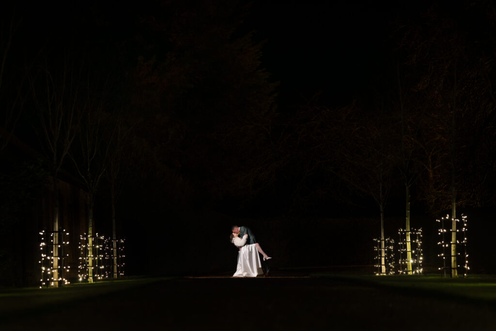 A couple stands in the center of a dark outdoor path at night, embraced in a romantic dip kiss, surrounded by trees wrapped in soft white fairy lights.