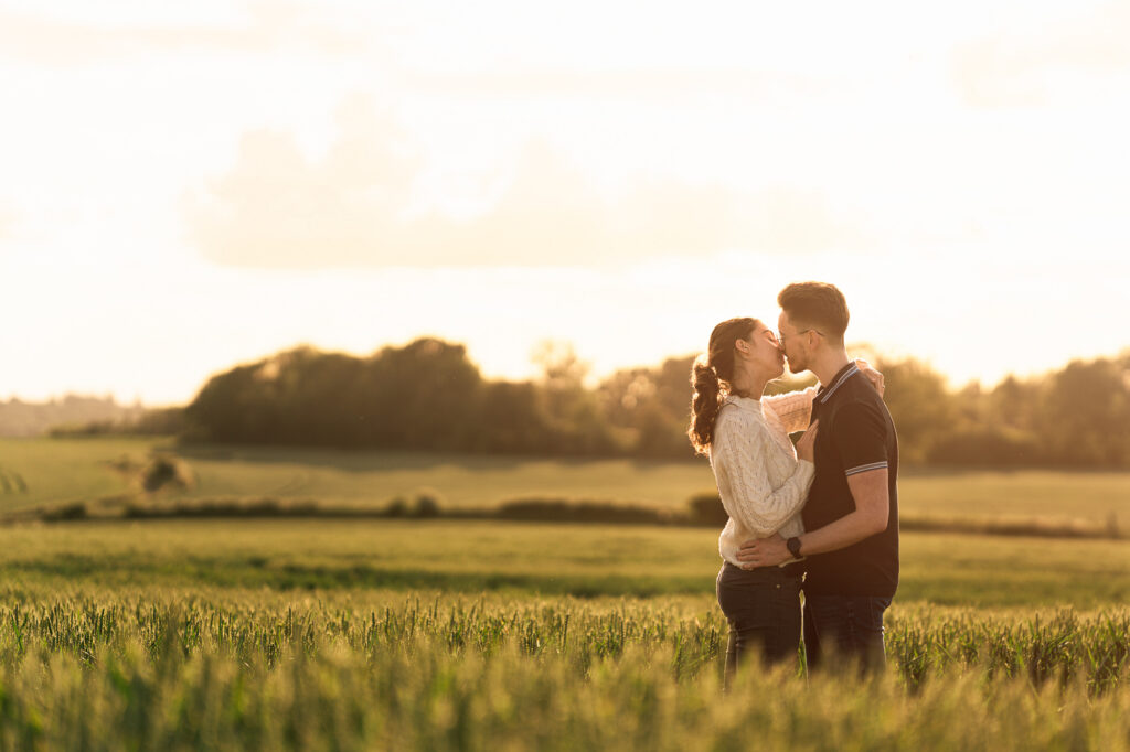 A couple embraces and shares a kiss in a sunlit field with green grass and trees in the background, creating a warm and romantic atmosphere at sunset.