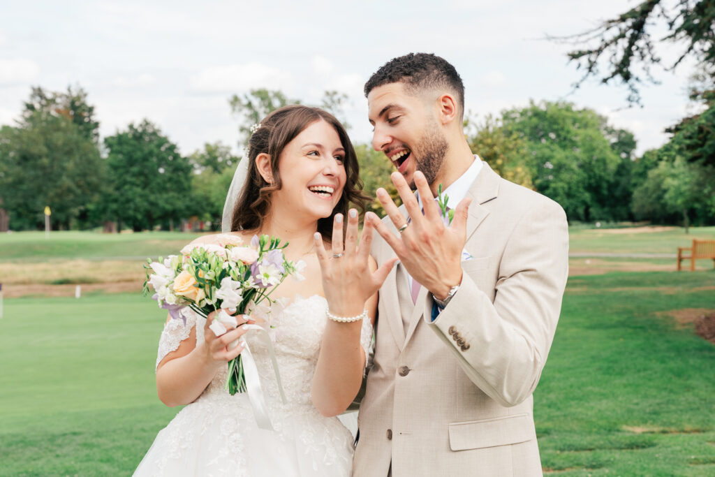 A joyful bride and groom stand outdoors on grass, smiling and showing their wedding rings. The bride holds a bouquet of flowers and wears a white dress, while the groom wears a beige suit. Green trees and a blue sky are in the background.