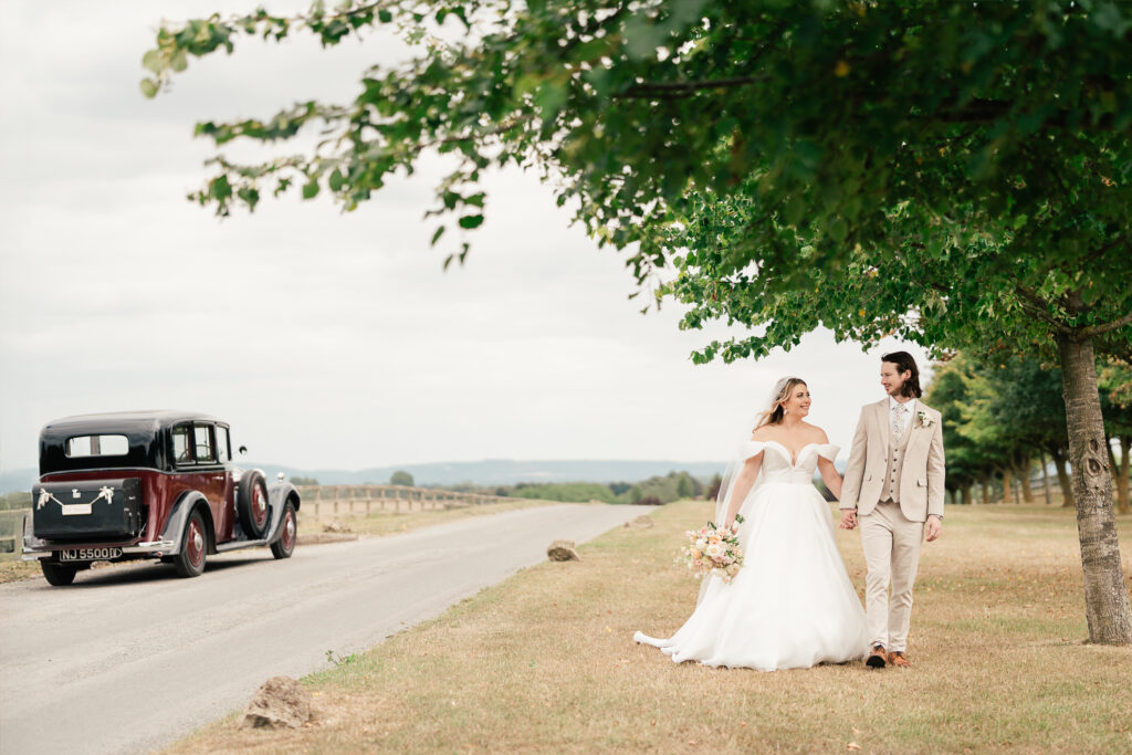 A bride and groom walk hand in hand on a grassy roadside under a leafy tree. The bride wears a white gown and holds a bouquet, while the groom is in a beige suit. A vintage car is parked on the nearby road.