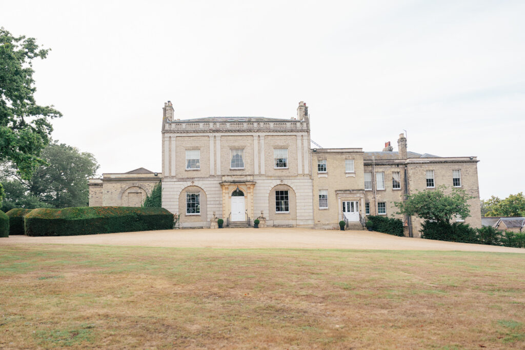 A large, historic, light-brick mansion with tall windows and white doors sits atop a gently sloping lawn with trimmed bushes and trees on each side under a cloudy sky.